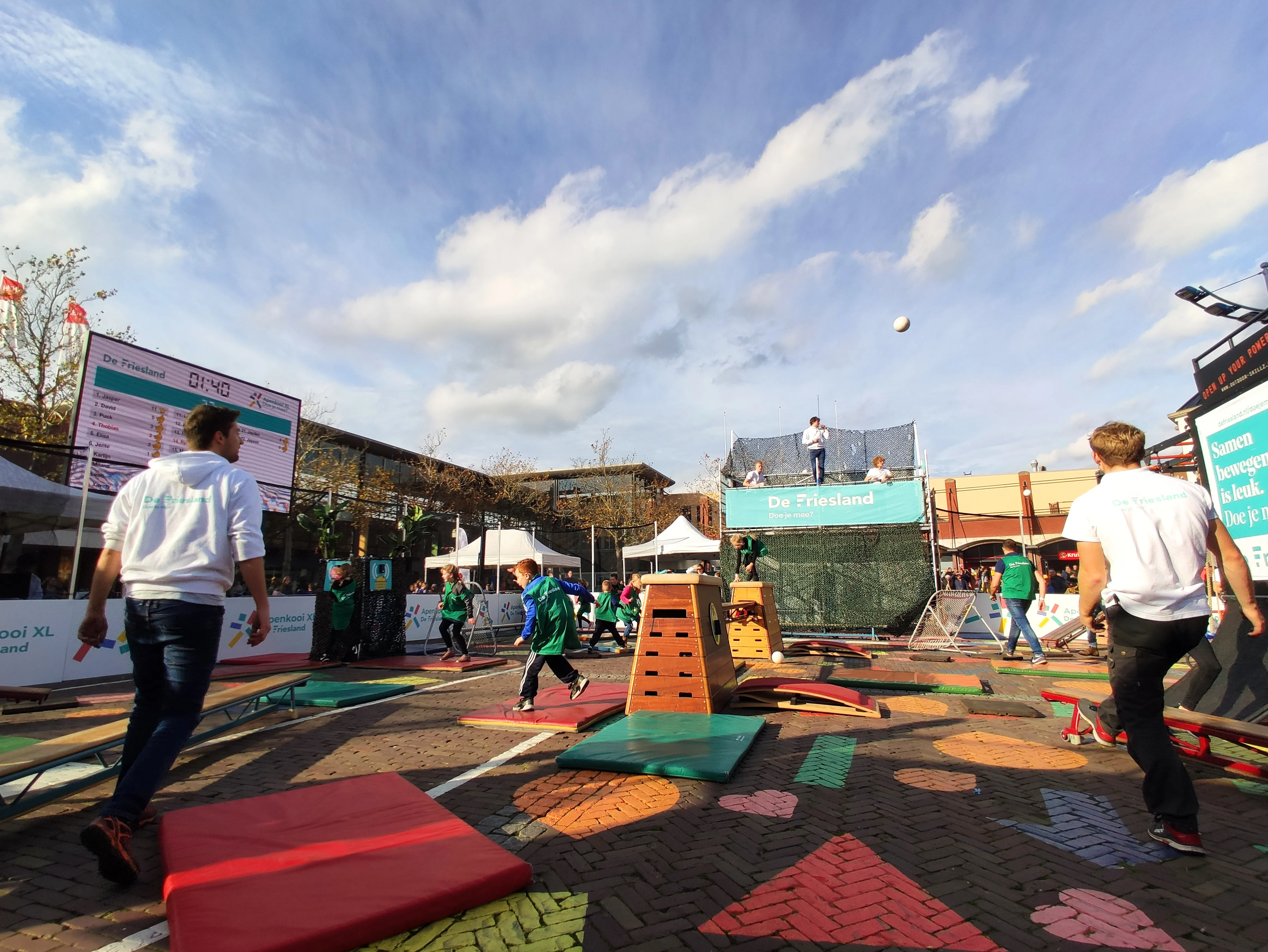 Kinderen spelen apenkooi in de mobiele ApenkooiXL-arena op een plein, met turntoestellen, matten en een groot live scorebord
